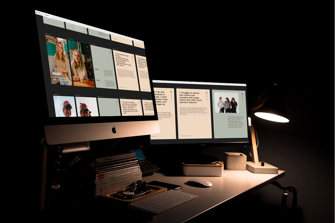 A dual-monitor studio workspace at night, one screen showing a website design and the other a document, lit by a desk lamp.